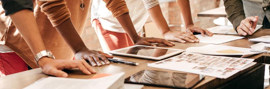 Several people leaning on a table full of papers and pads.